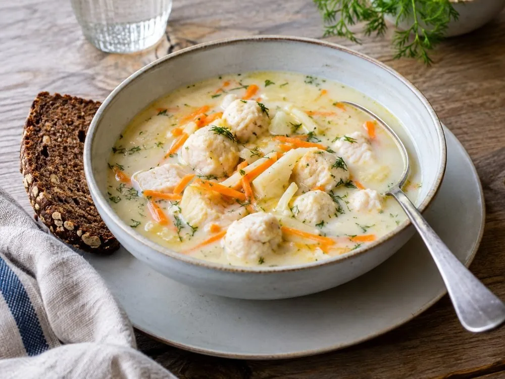 Eine Schüssel Bergen-Fischsuppe, hell und cremig, mit fein geraspelten Karotten und Wurzelgemüse, kleinen Fischklößchen und einem Löffel, der am Rand lehnt.