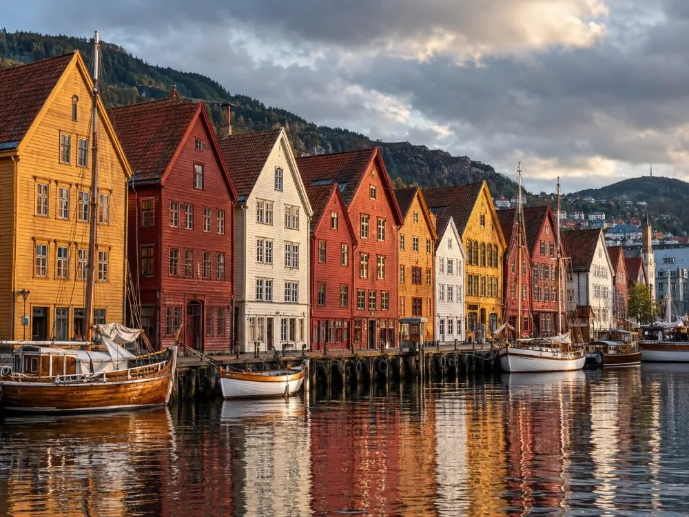 Die hölzernen Giebelfronten von Bryggen entlang des Hafens im tiefen Nachmittagslicht, ockerfarben, rot und creme, leicht geneigt, mit Booten, die davor vertäut sind.
